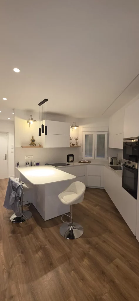 Piso reformado integralmente en León: cocina blanca abierta con isla, taburetes y suelo laminado de madera